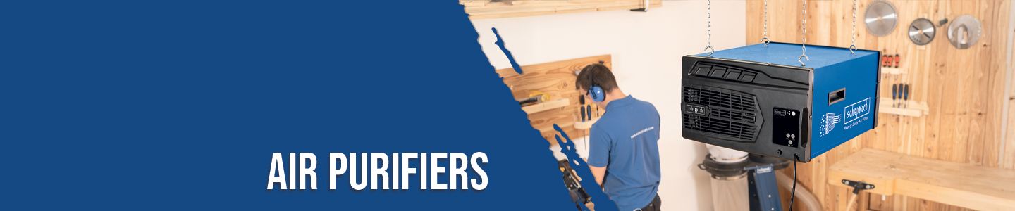 A worker in blue clothing operates an air purifier in a workshop. The air purifier is blue and suspended from the ceiling. Tools and wooden shelves are visible in the background.