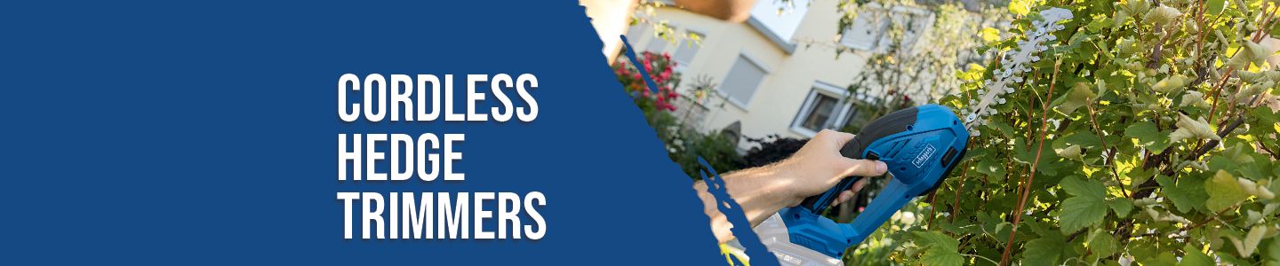 A Scheppach cordless hedge trimmer in use. One hand holds the handle while the other operates the trigger. A house with green plants is visible in the background.