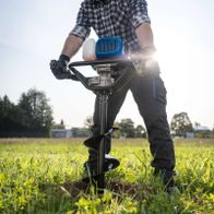 Person operating an earth auger tool in a field, showing the auger bit in use