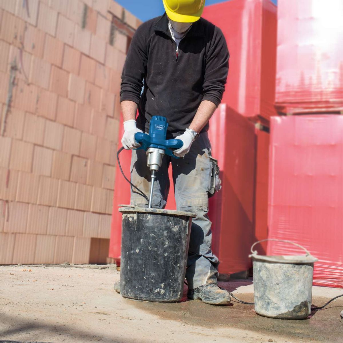 Scheppach paint and mortar stirrer with handle and tool holder, shown in use with protective gear and buckets.