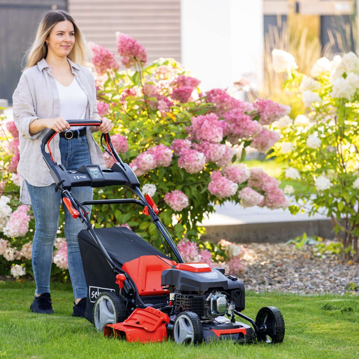 Person with Scheppach MS225-53E lawnmower beside flowering hedge, featuring self-propelled and E-start mechanism