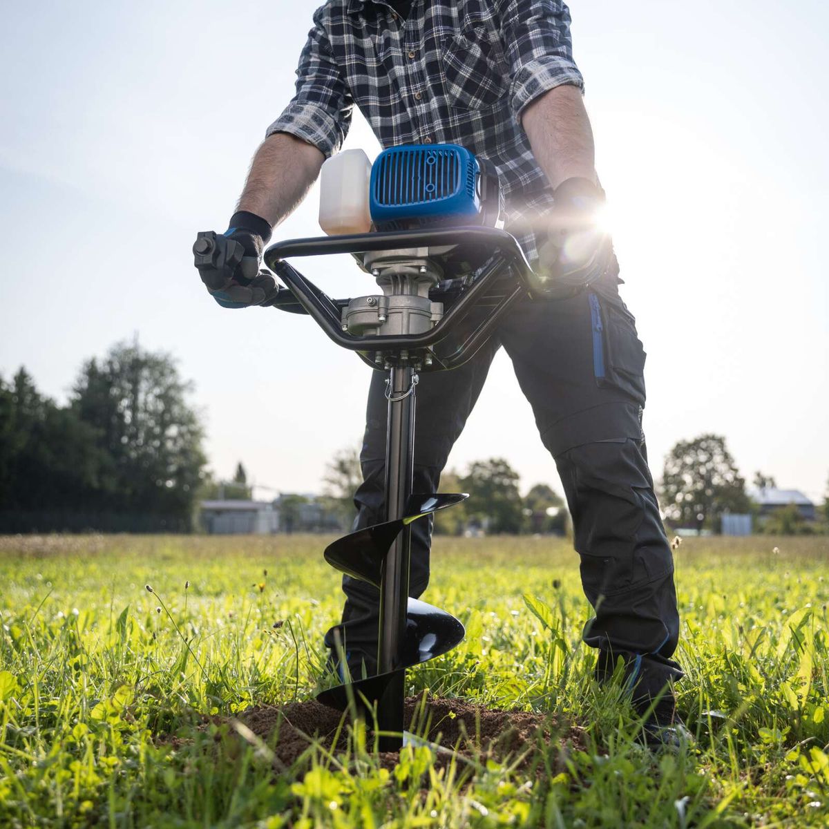 Person operating an earth auger tool in a field, showing the auger bit in use