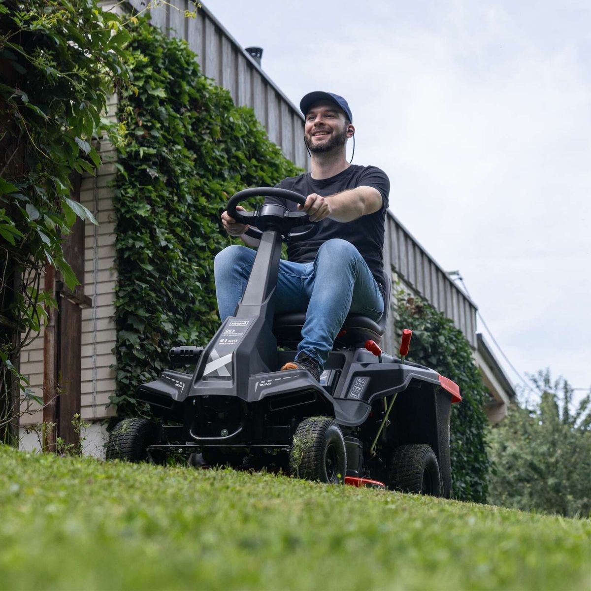 Person operating Scheppach ride-on lawnmower with LED lighting and cover, highlighting 61 cm cutting width and 150-litre bin