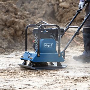 A Scheppach plate compactor in use on a construction site. The worker holds the handle of the machine firmly.