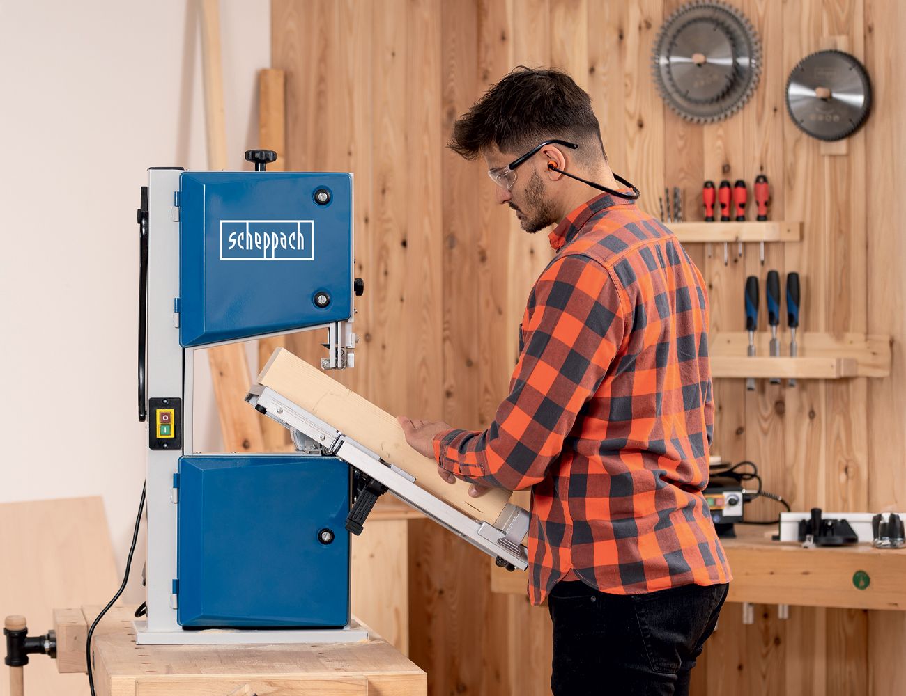 A man operates a Scheppach band saw in a workshop. Tools and wooden shelves are visible in the background.