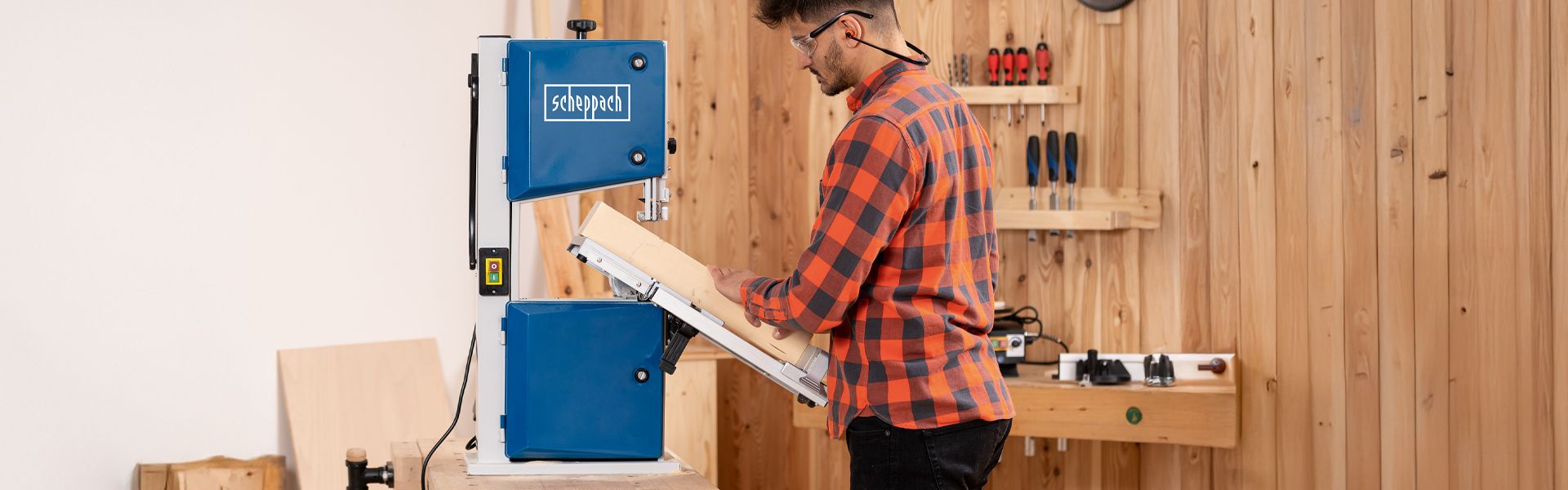 A man operates a Scheppach band saw in a workshop. Tools and wooden shelves are visible in the background.