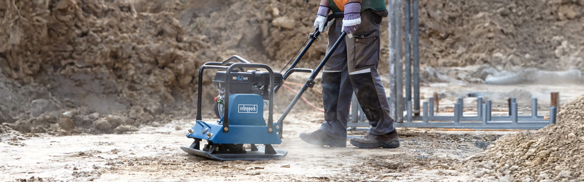 A worker operates a Scheppach vibrating plate on a construction site. In the background, scaffolding and a concrete wall are visible.